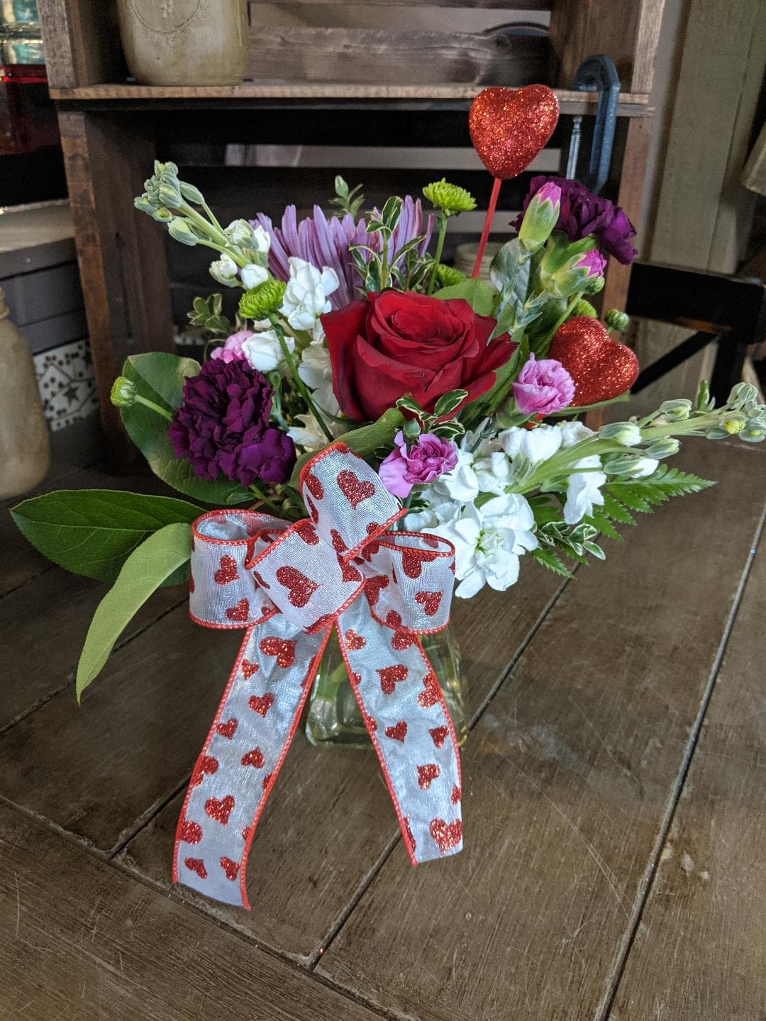 Kindest Heart Bouquet Custom Design by Cold Spring Floral - Kindest Heart Bouquet is arranged thoughtfully in a clear glass vase for that someone on your list that has the Kindest Heart.  A single Red Rose is paired with a Lavender Fuji Mum, White Stock, Deep Purple Carnations, Lavender miniature Carnations  and a Valentines ribbon.  All Flowers, Containers, vases and additions such as hearts, balloons and ribbons are subject to substitution based upon availability and will be coordinated beautifully. 
