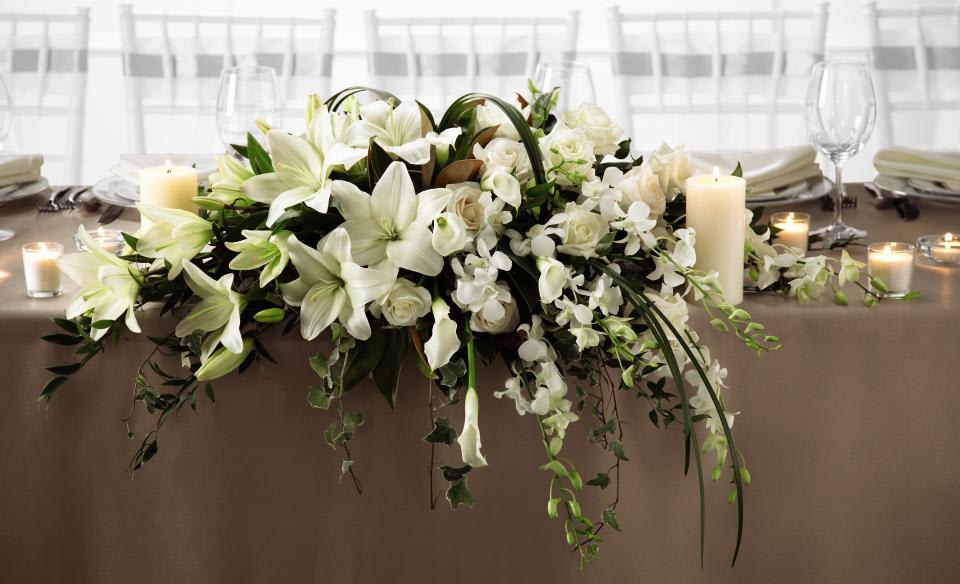 The White Linen Arrangement - The White Linen Arrangement speaks to the true nature of wedding  elegance. White Dendrobium Orchids, Asiatic Lilies, mini calla lilies,  and roses are accented with an assortment of lush greens and arranged to  add to the decor of the wedding party's table with its sweet  sophistication. Approx. 22âL x 40W x 18H 