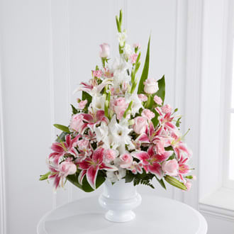The FTD Precious Peace Arrangement - The FTD® Precious Peace™ Arrangement is an exquisite display of serene wishes and grace. Soft pink roses Peruvian lilies and mini carnations are arranged amongst dazzling Stargazer lilies and white gladiolus gorgeously accented with lush greens. Perfectly situated in a white plastic designer urn this stunning arrangement will add to the beauty and elegance of their service or memorial.