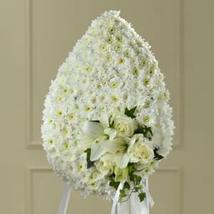 The FTD Solace Teardrop - You can be sure that this exquisite expression of sympathy crafted in the white of heavenly hope will make a moving and impressive addition to a wake funeral or burial service. Reflecting the form of a “teardrop ” this generously scaled spray constructed by a local FTD artisan florist includes white pompons roses and Asiatic lilies accented with a white satin ribbon bow. It comes on a metal stand to give it the ultimate flexibility for display at a funeral home in a church or at a cemetery.