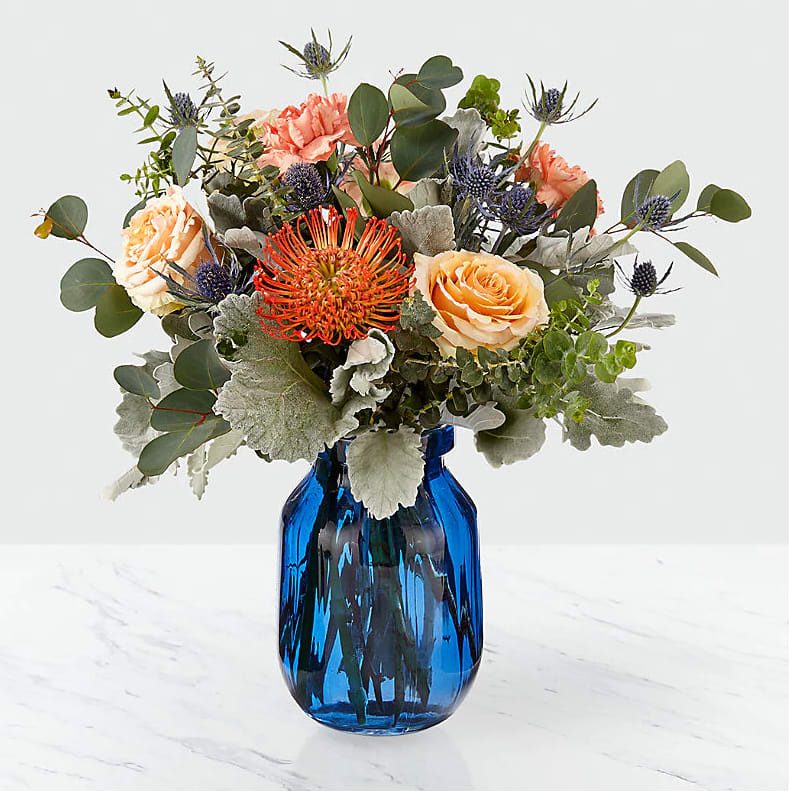 The FTD® Muse™ Bouquet The FTD® Muse™ Bouquet - There's just something about these soft, muted hues that will simply take your breath away. A dazzling combination of roses, carnations, and pincushion protea are handcrafted together within a bold blue glass vase. Accented with baby blue and silver dollar eucalyptus, this arrangement makes for a collection of flowers that is unlike any other. GOOD bouquet is approx. 15"H x 15"W.