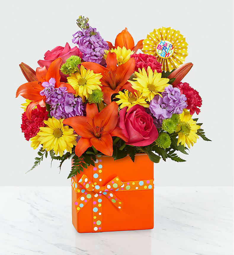 The FTD® Set to Celebrate™ Birthday Bouquet  - The Set to Celebrate™ Birthday Bouquet is ready to take their birthday celebration to the next level with a bright burst of color and perfectly arranged fresh blooms! Popping with personality and style, this flower bouquet brings together orange Asiatic Lilies, hot pink roses, lavender gilly flower, hot pink carnations, yellow traditional daisies, green button poms, and lush greens. Presented in a modern rectangular orange ceramic vase tied with a polka dotted orange ribbon to give it the look of a birthday present, this flower arrangement is then accented with a &quot;Happy Birthday&quot; pick to give it that ultimate party style. BETTER bouquet is approx. 16&quot;H x 13&quot;W.