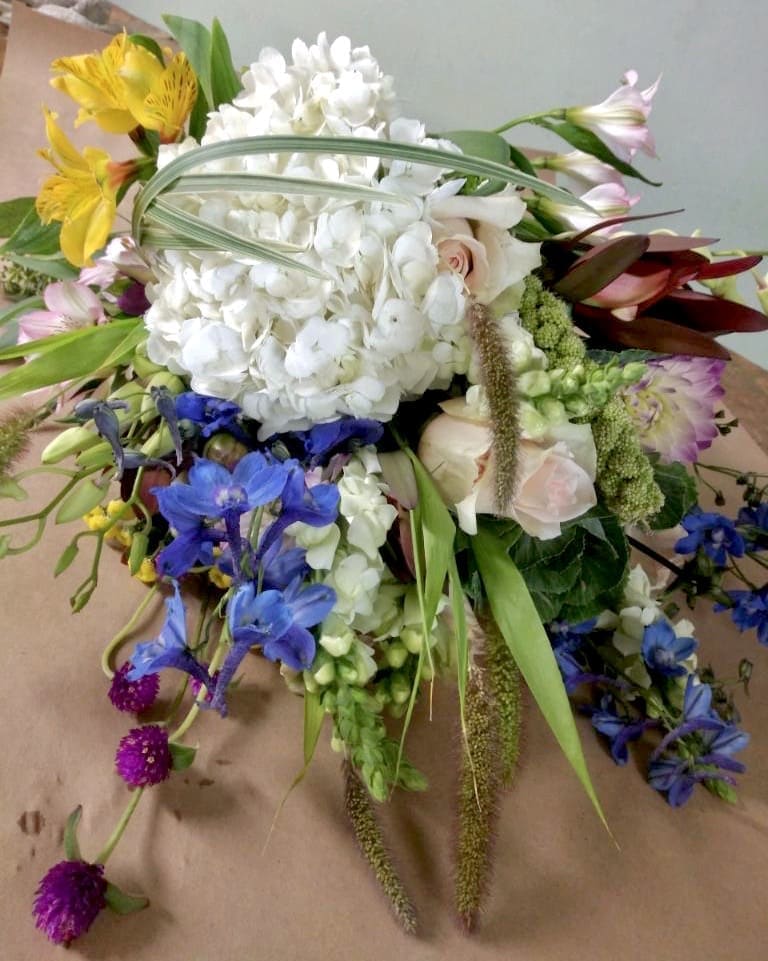 Rustic Wilderness Rustic Wilderness - A hand wrapped bouquet with white hydrangea, light pink roses, blue delphinium, safari sunset, and asiatic lily.