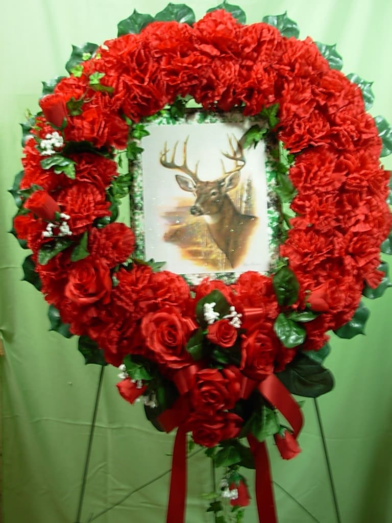 DEER HUNTER WREATH  NEW-S80 - OUR SILK DEER HUNTER WREATH  is made of SILK-PERMANENT red carnations and and with a cascading spray of red roses flowing down from the deer picture with red ribbon and comes on a tall easel stand..  The wreath is 27 inches wide and on the easel stands and impressive 56 inches tall.  Our silk and permanent flowers are an economical alternative to live fresh flowers and are also great for those last minute funeral orders when there is not enough time to create a fresh floral design.  Most all of our local cemeteries allow silk and permanent flowers to remain on the grave for some months.. Silk-permanent flowers are a very practical option during the freezing winter months or the extremely hot summer months in our area. 