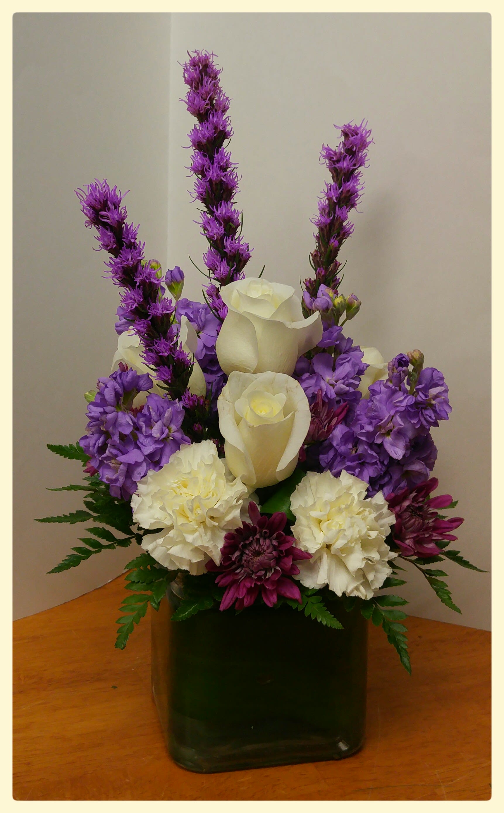 Purple Rain - Cream white roses, rich purple liatris, purple stock, white carnations and burgundy mums in a modern cube vase wrapped with a ti leaf...this elegant arrangement is sure to please!