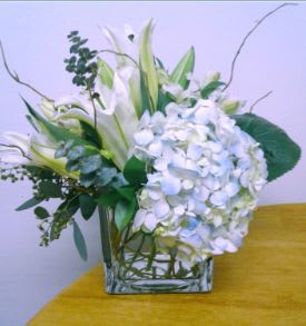 Above the Clouds! - This stunning arrangement incorporates white oriental lilies, white alstroemeria, textured greenery and curly willow, delightfully offset by a cloud of blue hydrangea.  Designed in a clear cube vase, this bouquet is the perfect way to say happy birthday, thank you, or thinking of you!