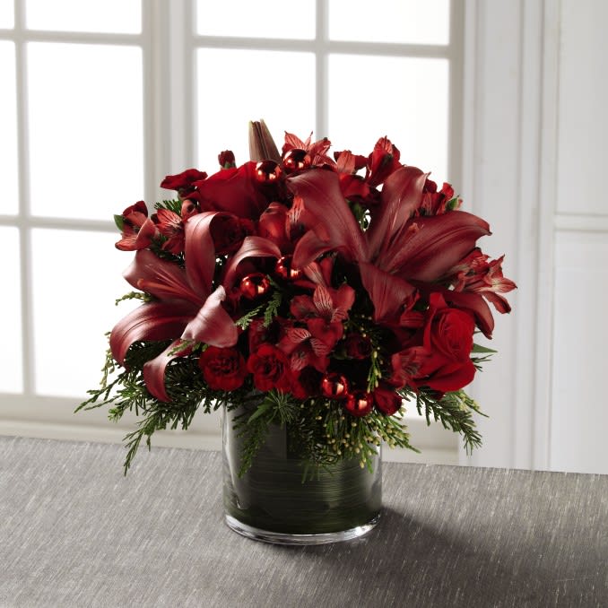 The FTD Season's Sparkle Bouquet - The holidays always seem to have an extra glimmering light set about those who make each moment count, adding love and friendship to an already joyous time. This fresh flower bouquet has been hand designed to reflect this treasured moment bringing together red roses, red Peruvian Lilies, red Asiatic Lilies and burgundy mini carnations, accented with an assortment of Christmas greens and shining red glass holiday balls. Presented in an oversized clear glass cylindrical vase lined elegantly with ti leaf material to give it a finished affect, this holiday flower bouquet is ready to make this yuletide season one to remember.