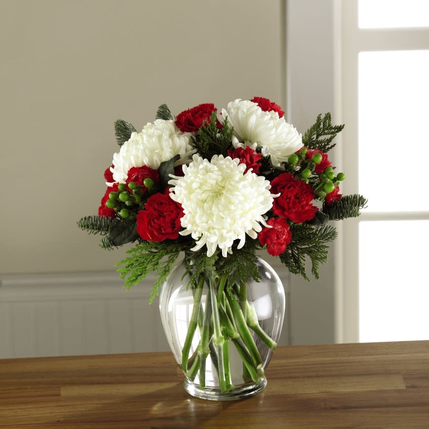 The FTD Holiday Enchantment Bouquet - Beautiful, bright, and blooming with a broad holiday appeal, this gorgeous Christmas flower bouquet is simply enchanting at every turn. Gorgeous and full white snowball chrysanthemums are not to be upstaged when arranged amongst rich red roses, red carnations, and red mini carnations, accented with red hypericum berries and assorted Christmas greens to create a standout yuletide display. Presented in an attractive clear glass vase, this gift of flower is ready to create a treasured holiday moment for your recipient. 