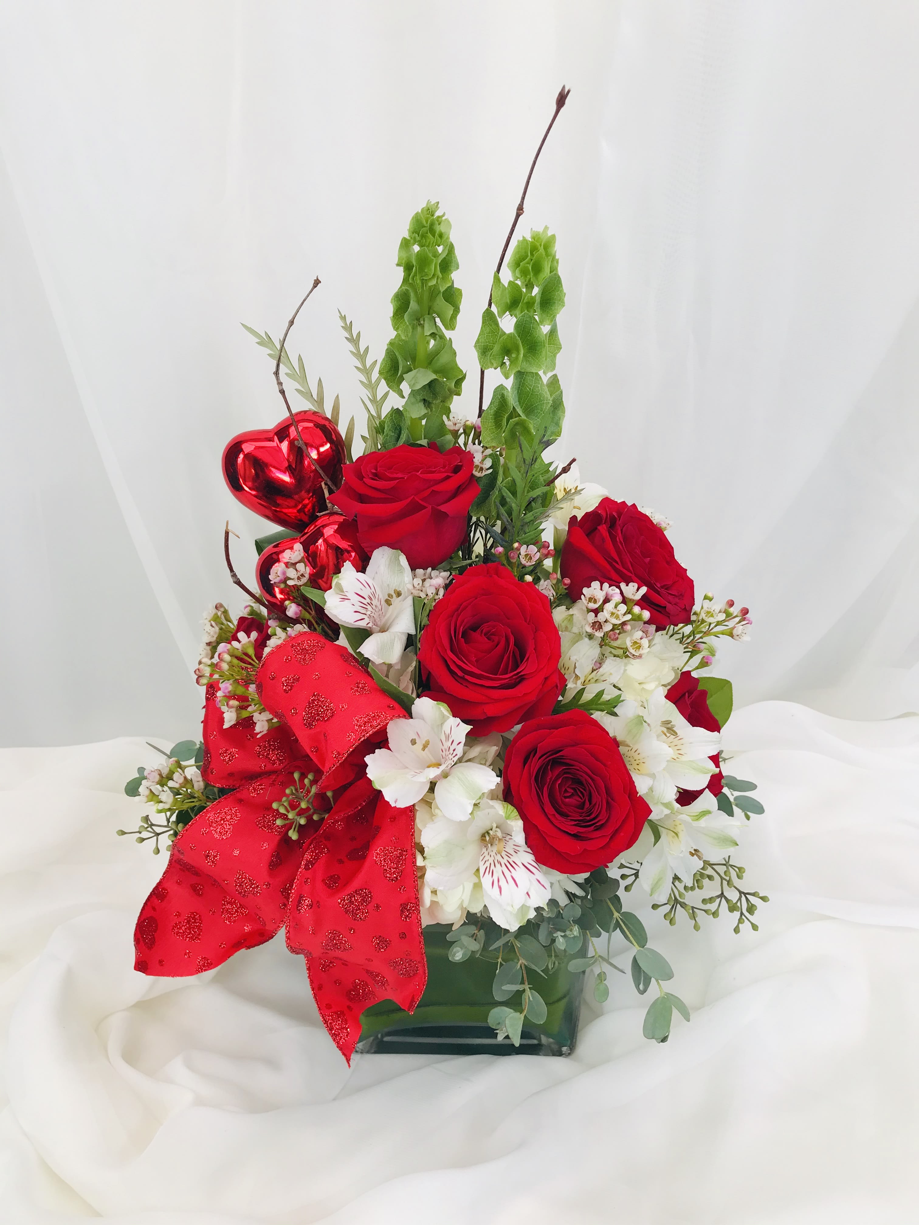 Pulling my Heart Strings Pulling my Heart Strings - This Valentine's Day arrangement is composed of red roses, bells of Ireland, alstroemerias, and a variety of greens. The piece contains two heart shaped accessories and a red bow. The arrangement sits in a square crystal vase.