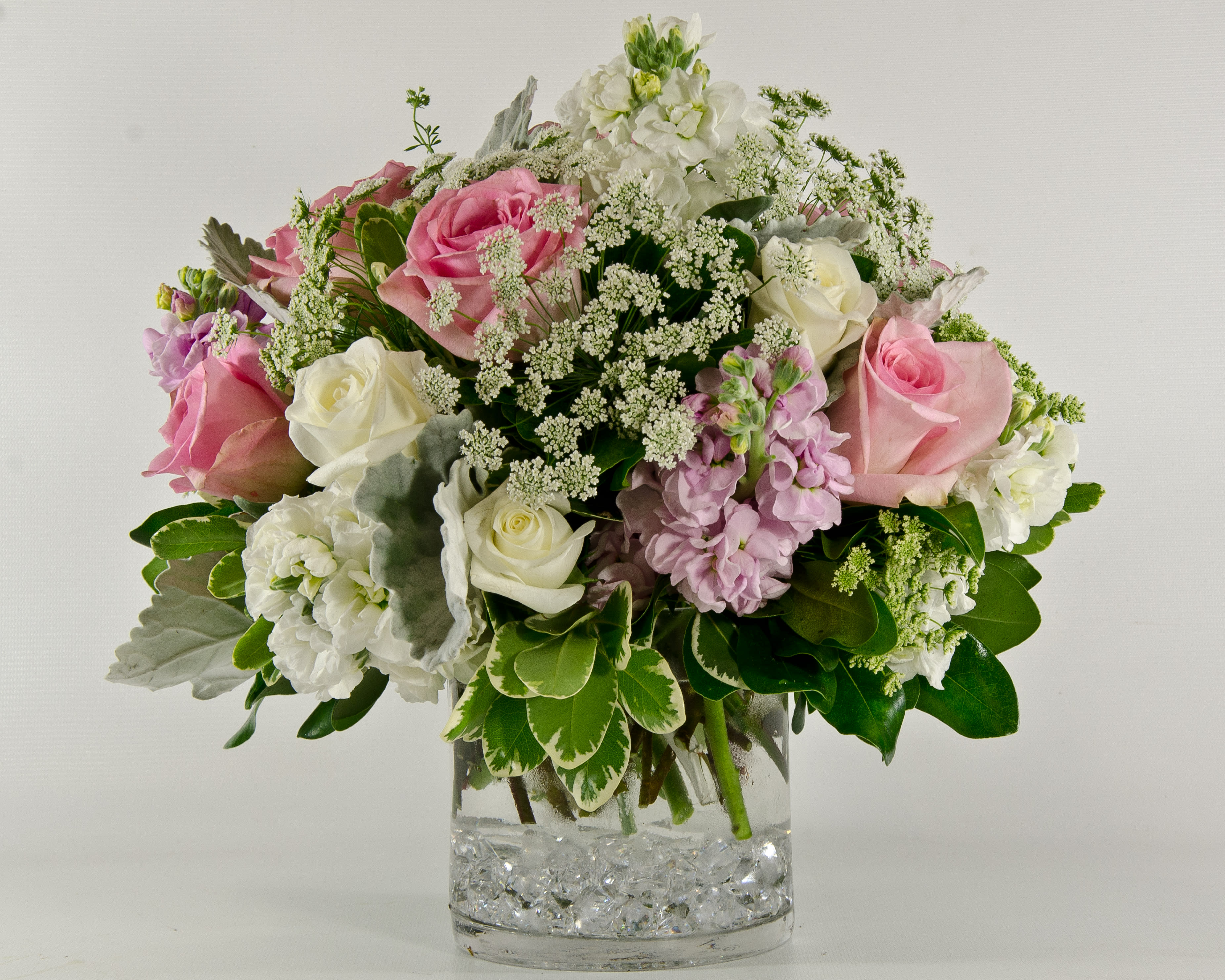 Beauty - A short compact table centerpiece with roses, stock,Queen Anne,dusty miller, mixed greenery design in a glass cylinder with glass bead accent.