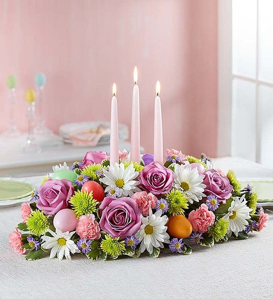 Easter Centerpiece Easter Centerpiece - Easter celebrations call for beautiful flowers. Our classic centerpiece is filled with pastel blooms and accented by colorful Easter eggs for a playful touch. Elegant taper candles finish off this charming arrangement, which is sure to be the bright spot of the holiday table. Hand-crafted centerpiece arrangement with lavender roses, Athos poms, white daisy poms, pink mini carnations and purple monte casino; accented with assorted greenery and colorful plastic Easter eggs