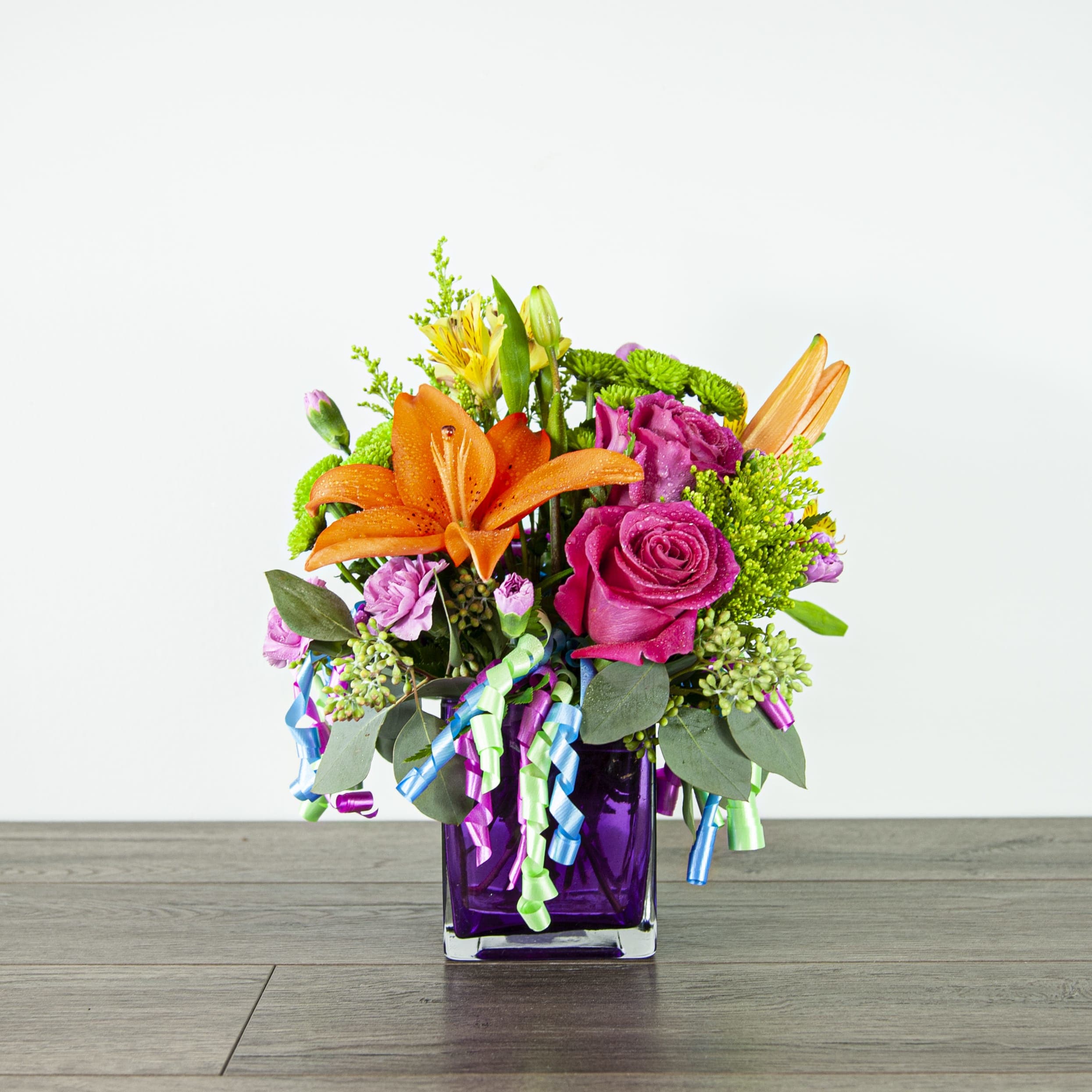 Birthday Party (EXP 59-B) - This colorful and festive centerpiece is ready for any party. The party look is completed with streamers in a violet cubed glass container.  Blooms in this arrangement: - orange lily, hot pink roses, green button poms, mini carnations, and alstromeria   