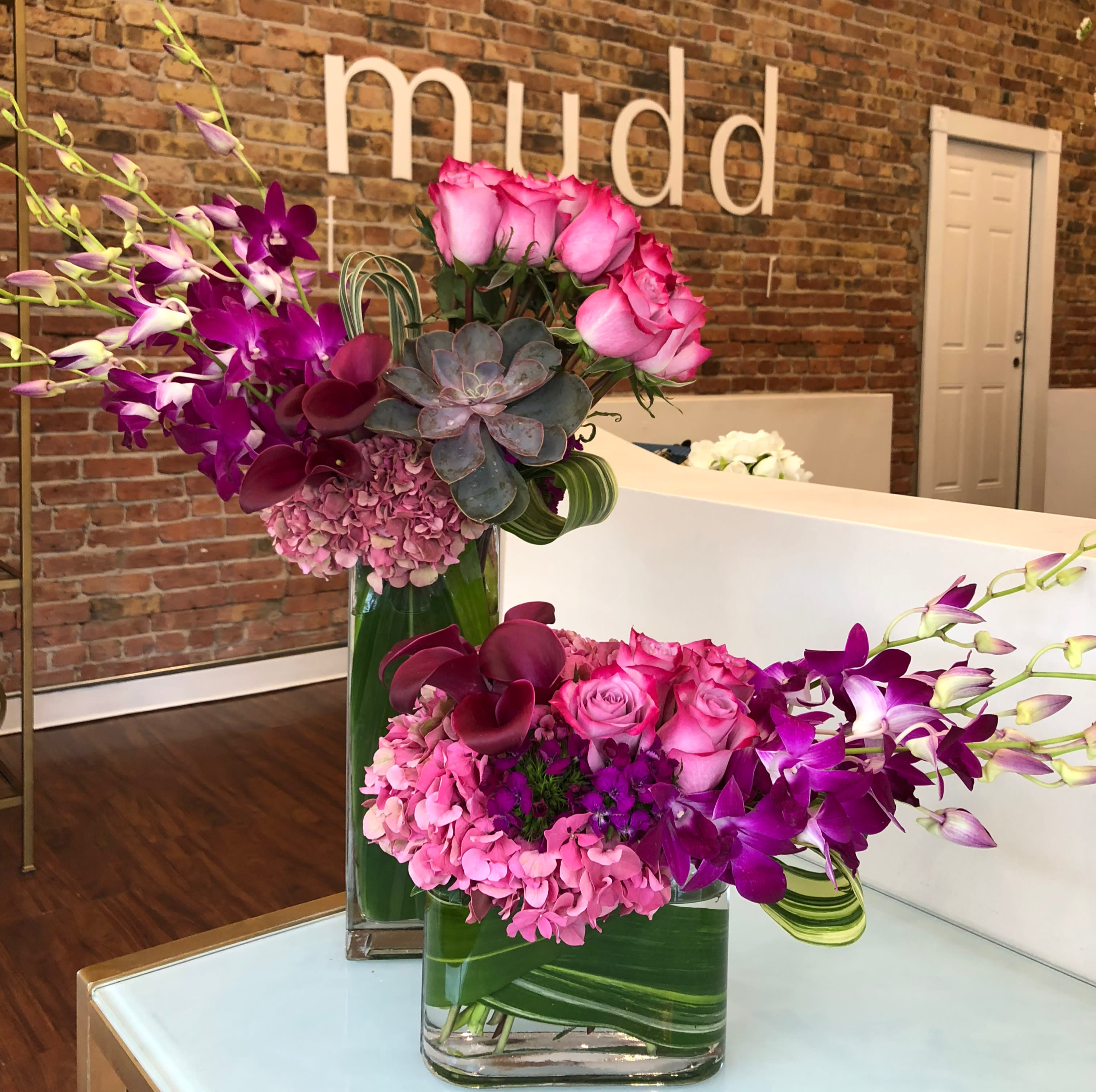 Signature Duo of Purple Flowers - Mudd Fleur signature two piece series of purple and pink flowers in clear glass rectangular vases lined with variegated leaf wraps. Flower varieties will vary on availability and will be in shades of purple, plum, lavender and pink. 