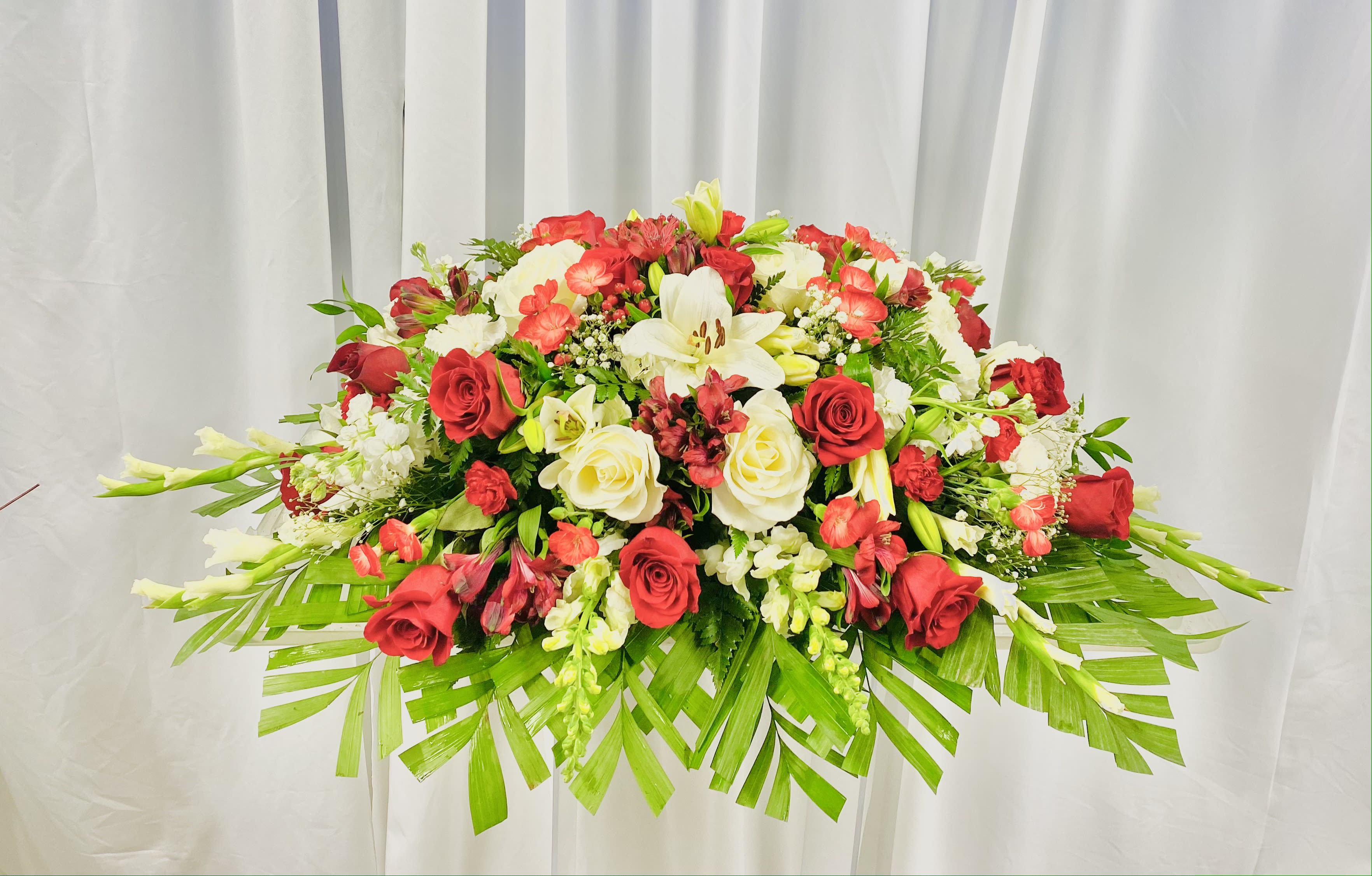 Red and White Serenity - A beautiful display of roses, lilies, snapdragon, altstromeria, and butterfly mums and more come together for a red and white half casket spray. Sized to fit both open and closed caskets. 