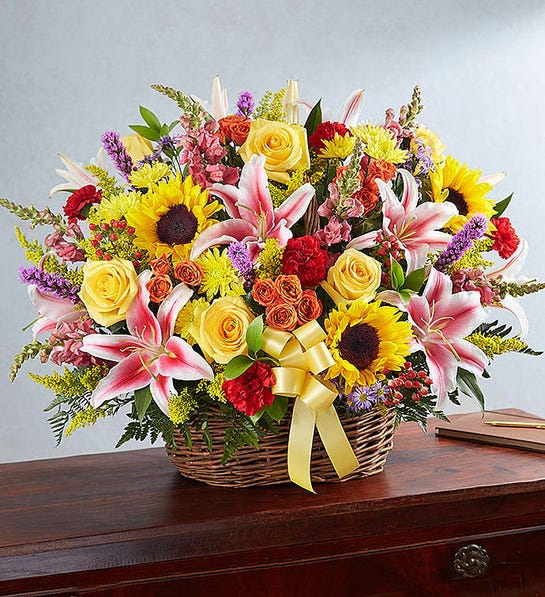 Multicolor Bright Sympathy Basket Arrangement - There’s a warmth and radiance to our bright basket arrangement, which beautifully captures the spirit of a departed loved one. Handcrafted by our expert florists, and filled with vibrant blooms, it is typically brought home after the services by a family member, bringing hope and light to them when they need it most.  Basket arrangement with red carnations and hypericum; orange spray roses; yellow roses, sunflowers and cushion poms; pink Stargazer lilies and snapdragons; purple liatrus and monte casino; accented by soft, lush greenery and yellow bow  Appropriate for the funeral home Arrangement measures approximately 28&quot;H x 24&quot;L Our florists use only the freshest flowers available, so colors and varieties may vary