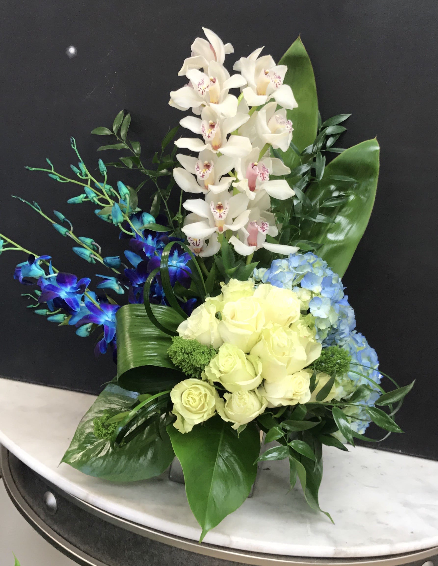 Step with me - White cymbidium and blue dendrobiums arranged in a silver vase with white roses, blue hyndrangea and greenery to show that person how your are feeling.. Perfect arrangement for every occasion. 
