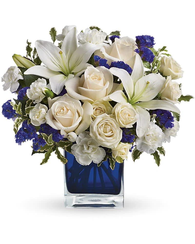 Blue Dreams - Send someone a bit of heaven with this beautiful bouquet. Luxurious crème roses and pure white lilies paint a peaceful picture inside a sapphire blue cube.  Crème roses, white asiatic lilies and white miniature carnations are mixed with bursts of purple statice and green pitta negra. Delivered in a glass Cube.