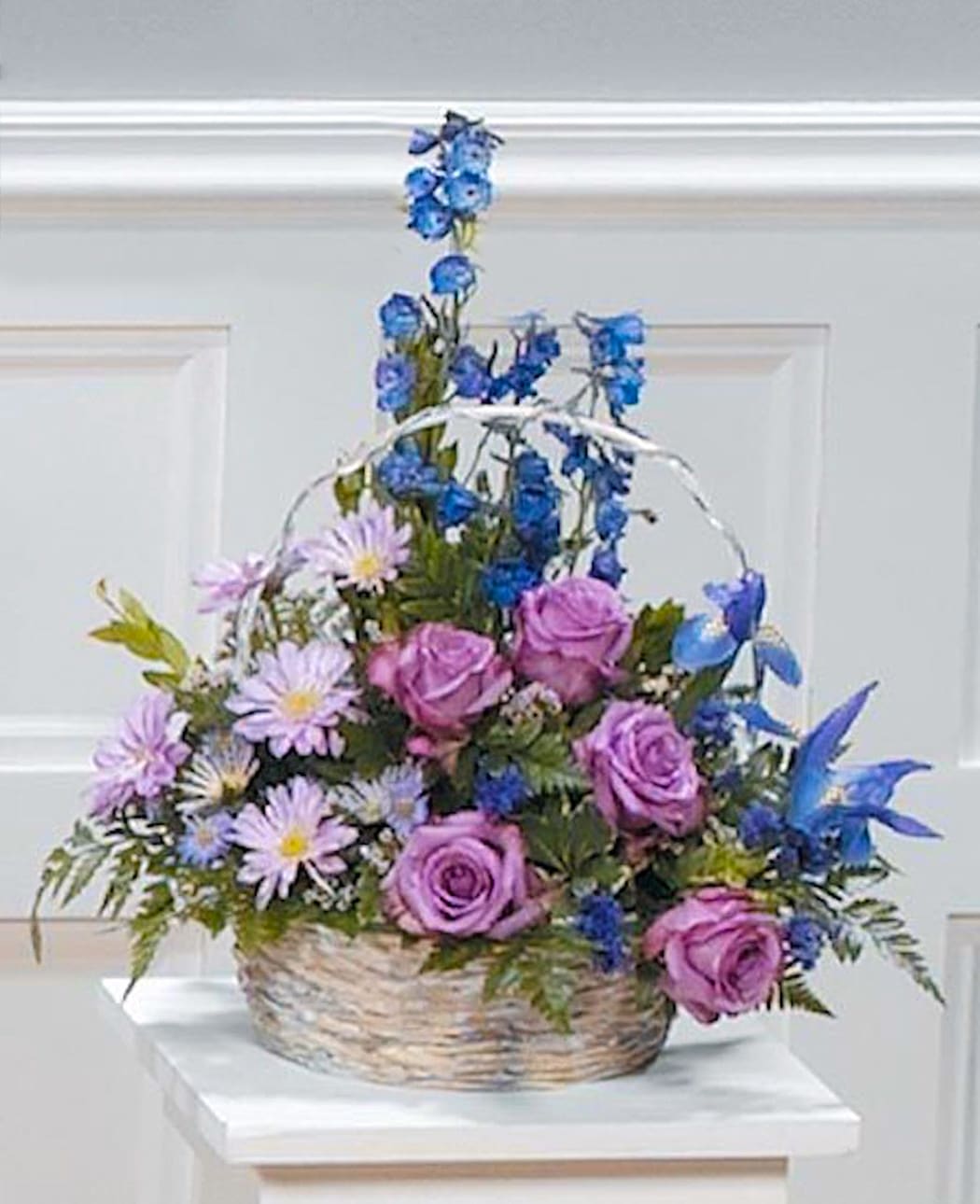 Serene Sentiment - A whitewash field basket holds a gathering of serene Lavendar Roses, Iris, Delphinium, Daisy Pompons and other select florals and greenery.
