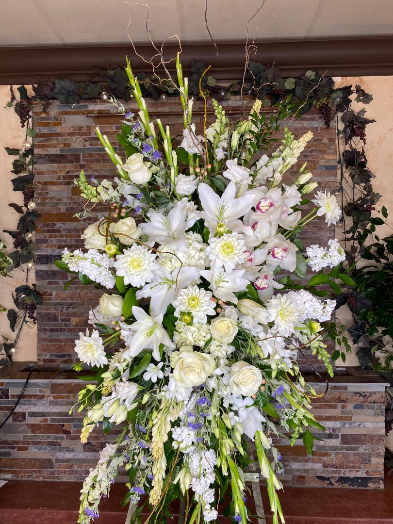 Heavens Grace Heavens Grace - This standing spray reaches over 8 feet in height, filled to the brim with white blooms including gladiolus, lilies, stock, roses and a stunning cymbidium orchid. Soft pastels are woven between the white to create finish off a beautiful design.