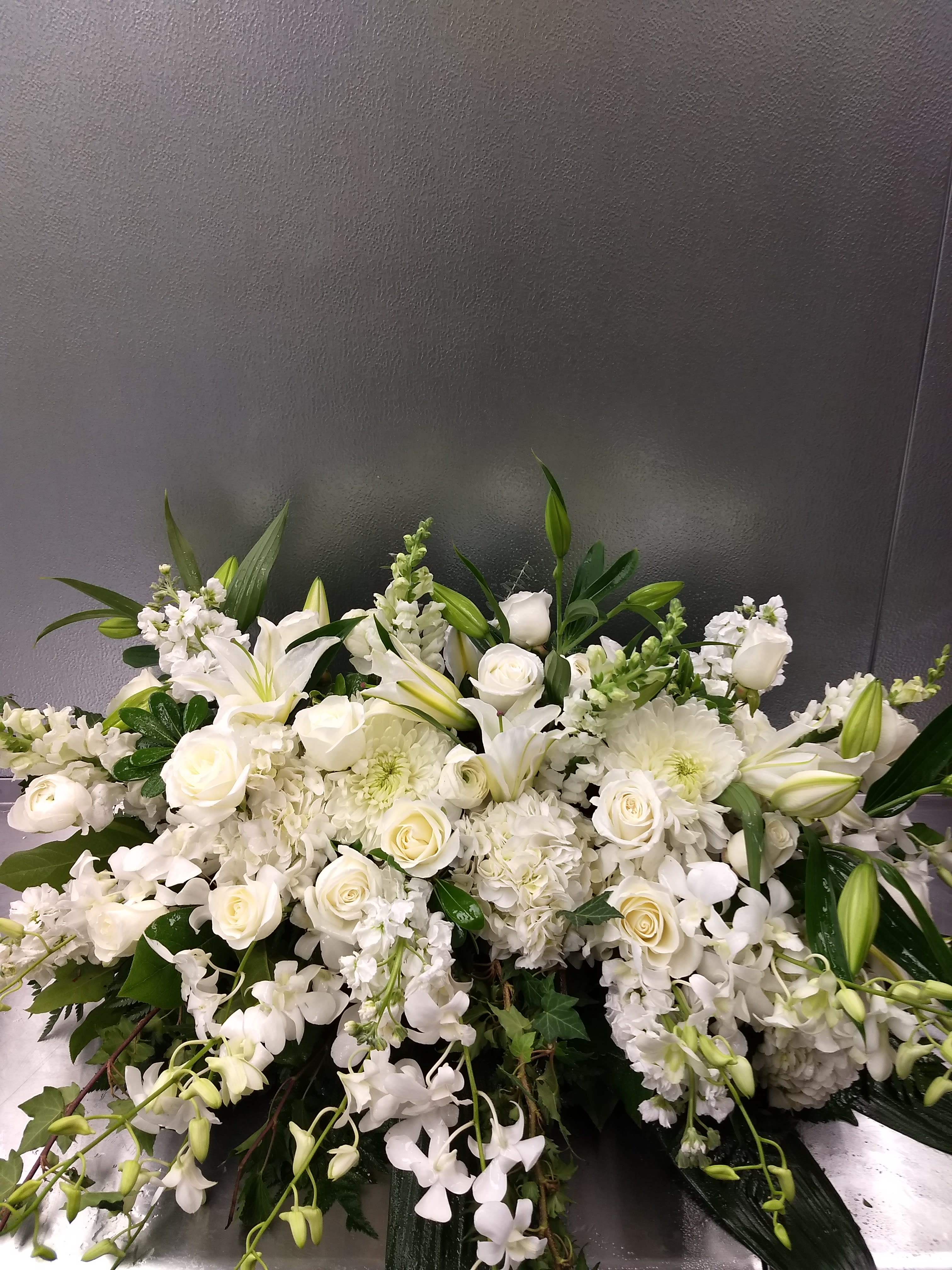 Elegant White Casket Spray - This elegant all white Casket Spray is overflowing with beautiful fragrant Lilies, Roses, orchids, Stock, and Snapdragons and more. This spray will fit on an open casket as well as when the casket is closed.  **** Please note: 48 hour notice MAYBE REQUIRED We custom-design this arrangement using best-of-day seasonal flowers. This image is a general representation of its size and style and may not feature the exact flowers shown. Due to disruptions in the global supply chain and staff shortages, we may have to make last minute substitutions to your selected design. If we need to make significant changes we will call you first to get your approval*** Thank you for understanding.