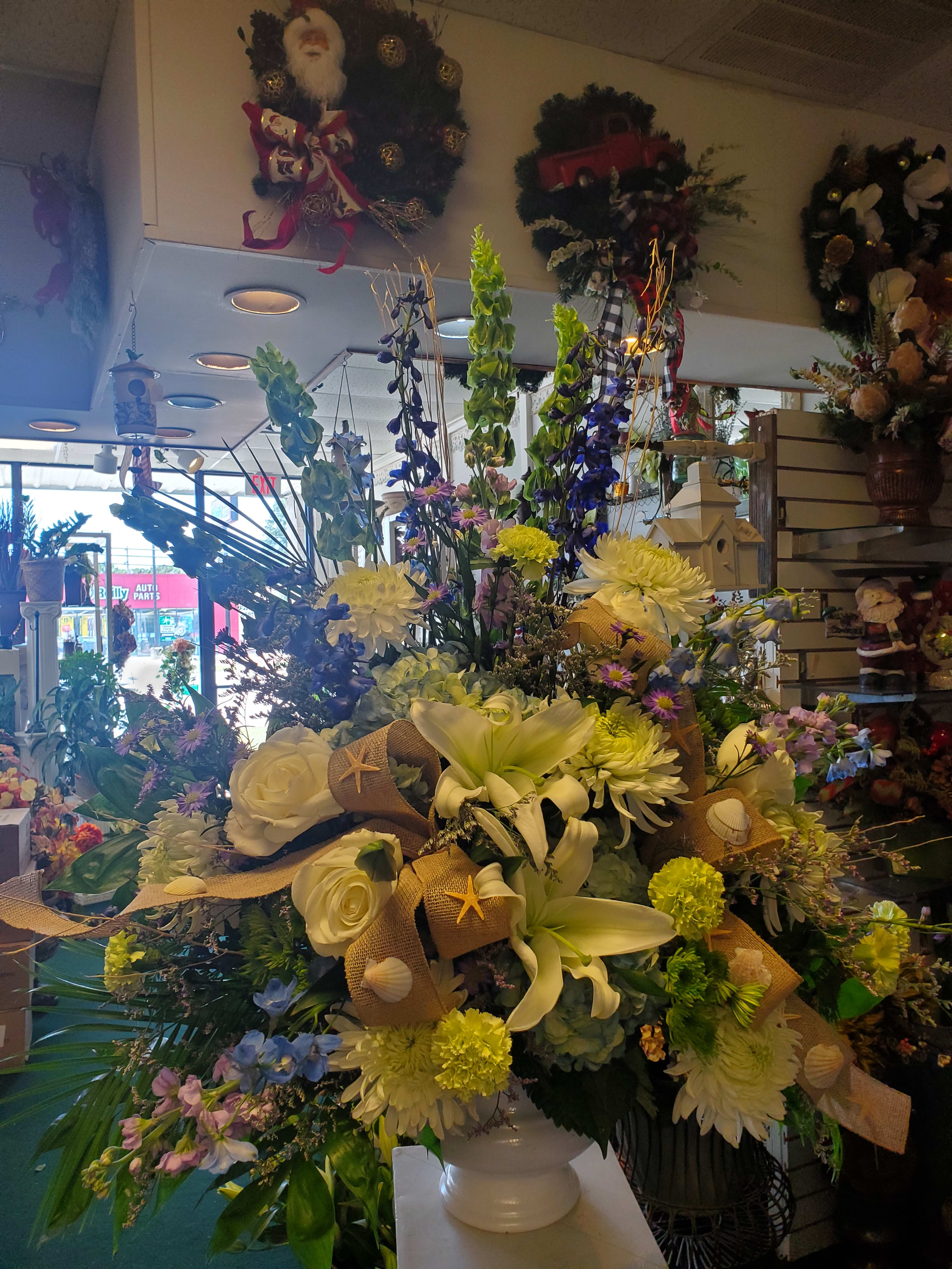 BEACH TRIBUTE URN - An urn arrangement featuring blue, white and green flowers, such as roses delphinum bells of ireland and lilys with accent s of ribbon and sea shells