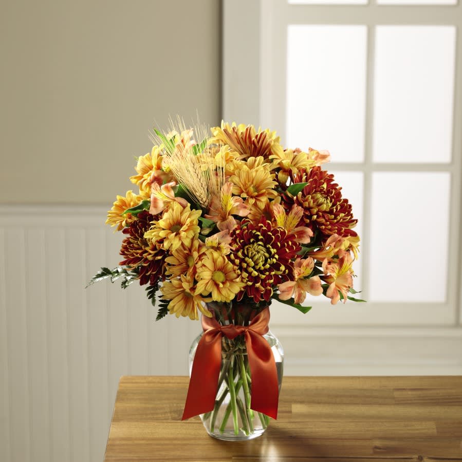 The FTD Autumn Roads Bouquet - Leading to a journey full of crisp fresh air and harvest sunlight, this stunning fall bouquet weaves together the fabric of this festive season. Bronze chrysanthemums, orange Peruvian Lilies, and butterscotch daisies blend together to create a warm and inviting bouquet accented with lush greens and dried wheat stems to give it a homespun, natural look. Presented in classic clear glass vase tied with a copper satin ribbon at the neck, this autumn flower arrangement is set to create a memorable thank you, thinking of you, or Thanksgiving gift.