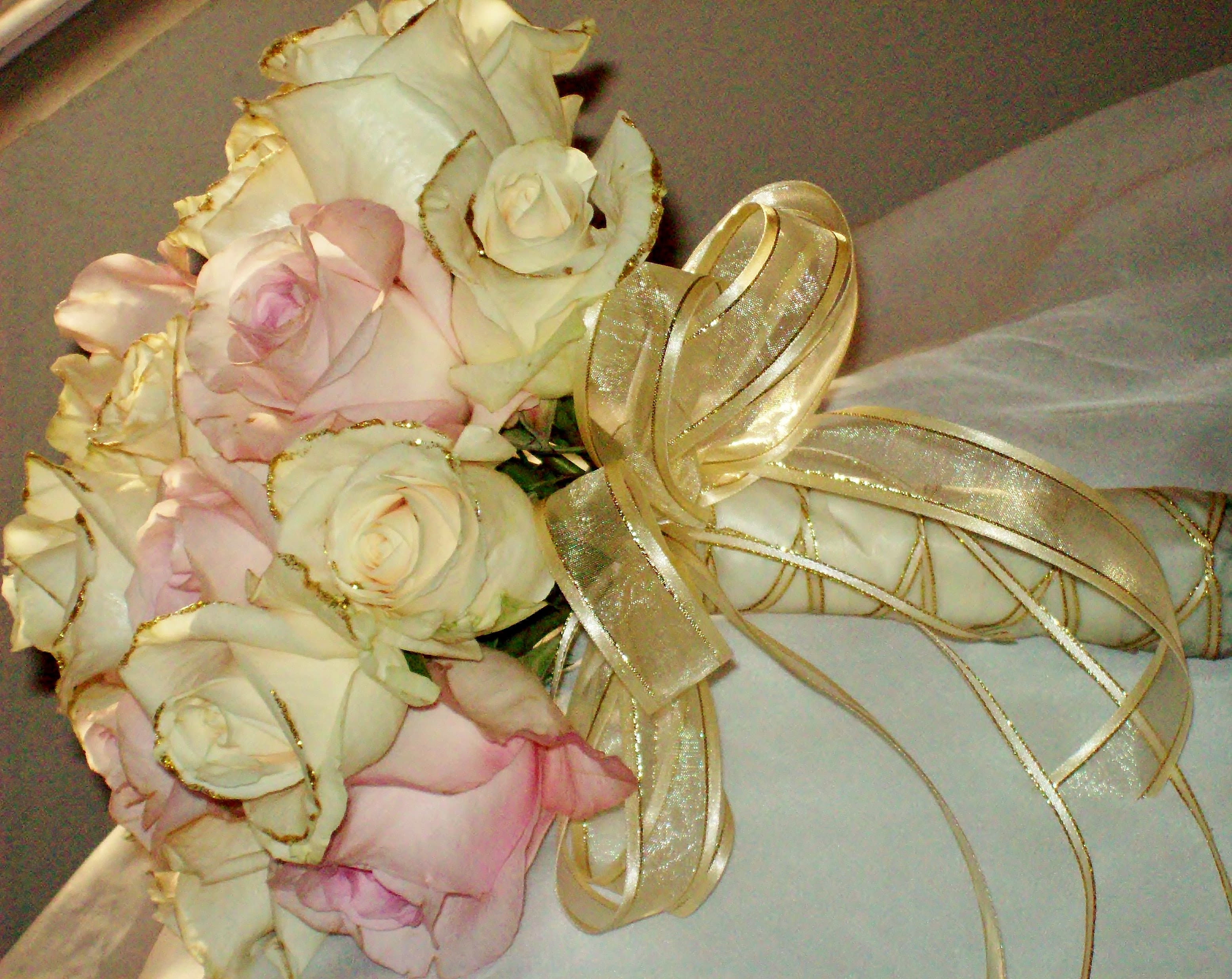 Pink & Gold Bridal Bouquet Pink & Gold Bridal Bouquet - A hand tied bouquet composed of Pink and Cream Roses tied in gold accented ribbon. Cream roses are accented with gold glitter and the Satin wrapped handle is decoratively accented with ribbon tails and a pearl pin line. This design measures approx. 12"W x 13"L. All bouquets must be ordered a minimum of 72 hours in advance.