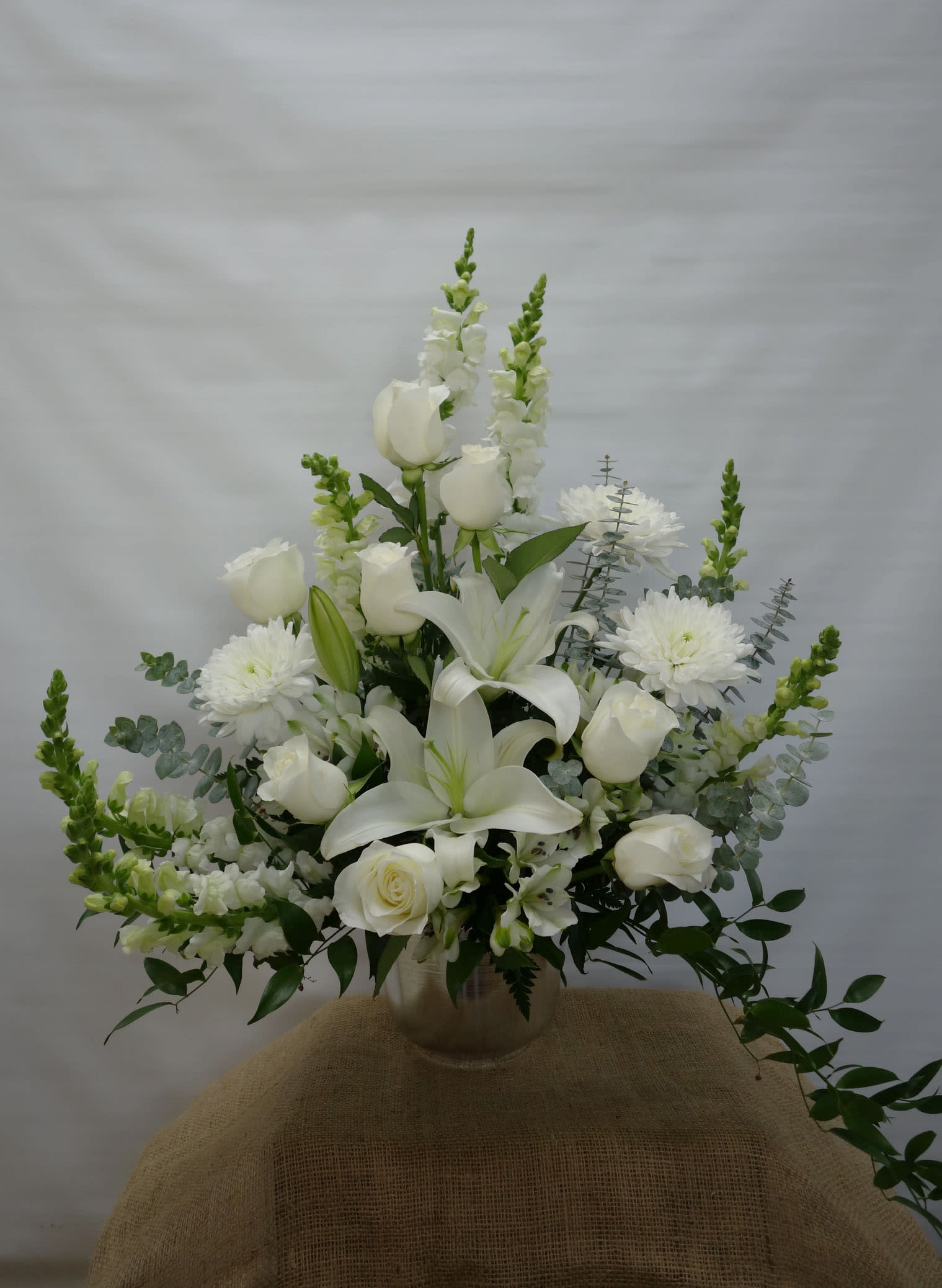 FB#05 Peaceful Reflections Basket - An all white arrangement featuring roses, lilies, spider mums, and filling greens in a silver jardiniere vase will be a lovely addition to the service.  The arrangement is approximately 24&quot; H and 24&quot; W.