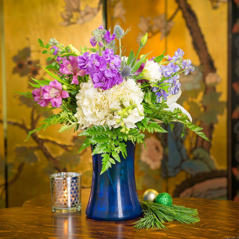LAVENDER ICICLE - POPS OF COLORFUL STOCK, WHITE HYDRANGEAS, AND BLUE THISTLE ALL GATHERED BY A BEAUTIFUL BLUE VASE.