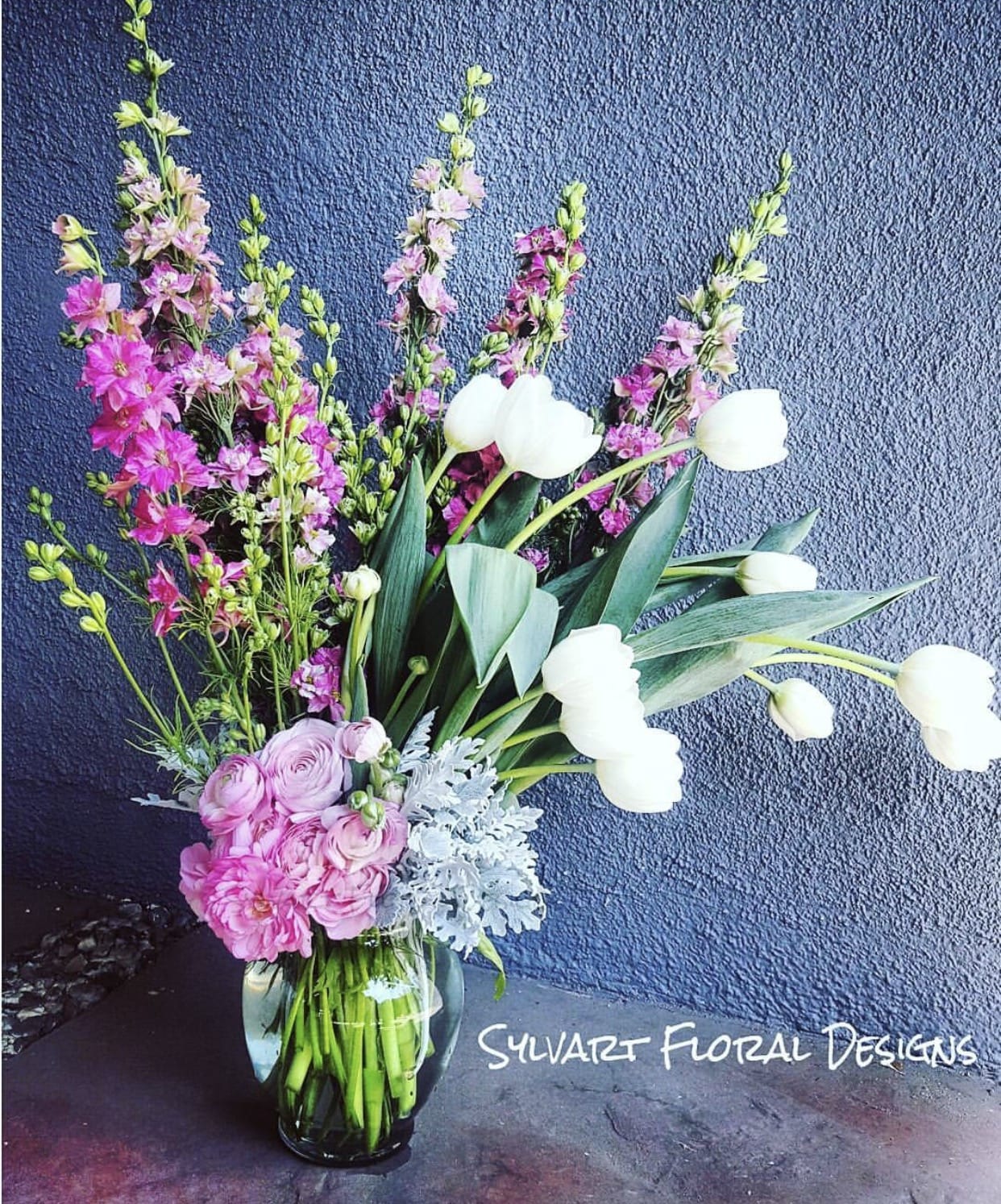 Flirty Tulips Flirty Tulips - This beautiful piece includes flirty tulips, dusty miller, anemone and a pink flair. (Florals and container might vary due to availability.) Note: If you would like to change floral color, please add your request in the note section when placing your order.