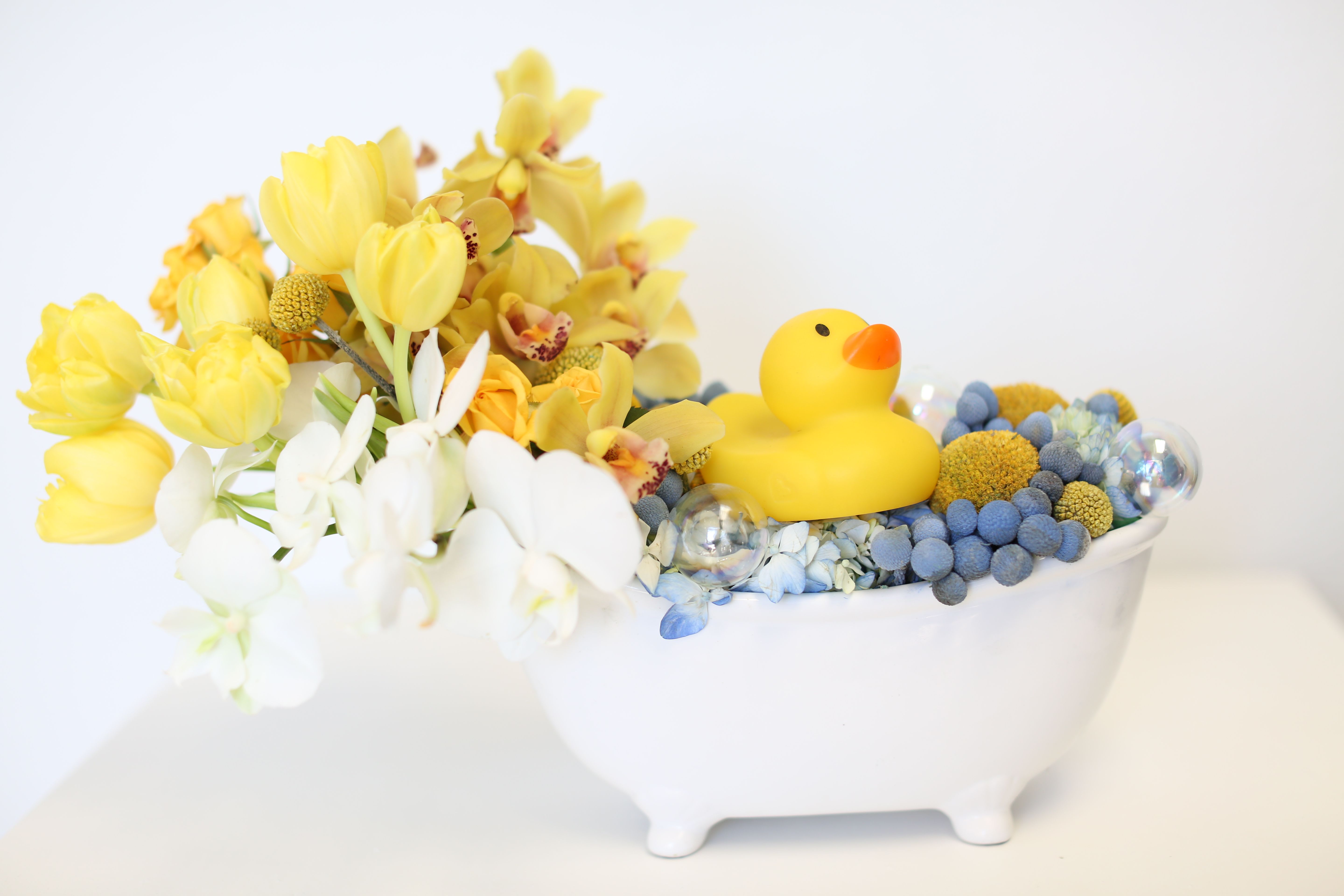 Dexter - This playful piece is perfect for sending to any new parents welcoming any a new child, or maybe for a child having their first birthday.  It is a 10&quot; wide ceramic bathtub with  Dexter, the rubber ducky, swimming in blue hydrangea and bubbles made of yellow billy balls and brazilin balls.  What fun is a bath without a little splash? Yellow mini cymbidium orchids, yellow tulips, and white mini Phalaenopsis orchids are spilling out to finish off this playful piece.  This piece is approximately 14&quot; wide and 10&quot; tall