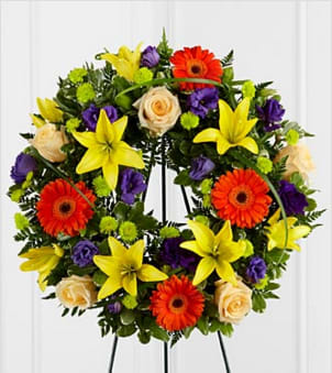 The FTD® Radiant Remembrance™ Wreath - The FTD® Radiant Remembrance™ Wreath is a brilliant burst of color and light that honors a life full of joy and beauty. Crème de la Crème roses create a symbol of peace arranged amongst the vibrant hues of purple lisianthus, orange gerbera daisies, orange Asiatic lilies, green button poms, and a variety of lush greens, forming a wonderful representation of a life well-lived. Displayed on a wire easel. Approximately 22-inches in diameter. Your purchase includes a complimentary personalized gift message.