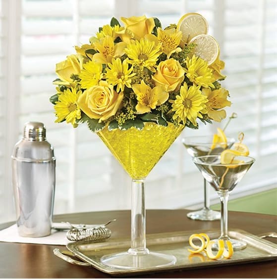 Lemon Drop - Send sunny sophistication with our truly original Happy Hour bouquet, inspired by the famous Lemon Drop Martini. Our floral version of the cosmopolitan cocktail is hand-crafted inside an oversized acrylic martini glass from cheerful yellow roses, daisies, alstroemeria and solidago. Topped with a faux lemon slice garnish, it's enough to fill anyone with sunshine and smiles.