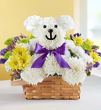 Beary Cheerful Basket - Here’s a cheerful surprise for loved ones of all ages! Handcrafted from fluffy white mums and crisp carnations, our cute and cuddly cub arrives in a charming handled basket filled with a vibrant mix of yellow daisy poms and purple statice. Holding a bouquet of yellow daisy poms in his paw and finished off with a purple satin bow, our truly original bear is a sweet way to show how much you care!