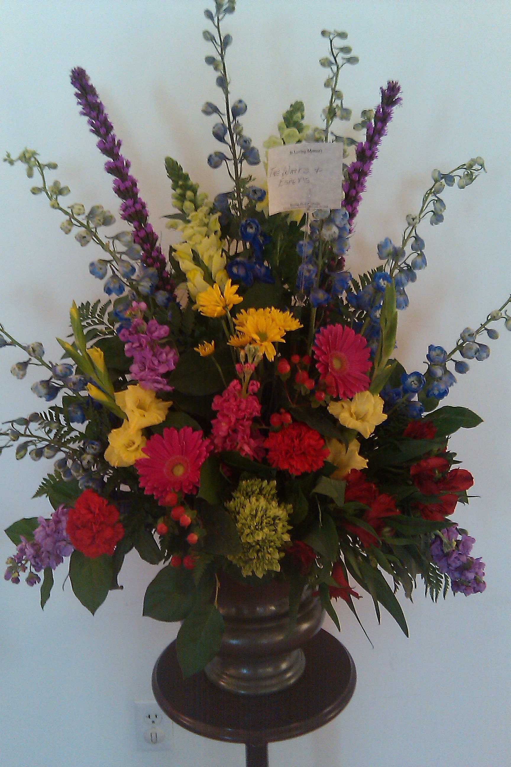  Comfort and Peace Arrangement - A Bright Garden Style table arrangement lends comfort and peaceful thoughts in this funeral design. Liatris, Gerber Daisy, Stock, Delphinium, Daisy Mums, Snap Dragon, Alstromeria, and Carnations are featured in this design.  Flower varieties and container may vary depending on availability and season.
