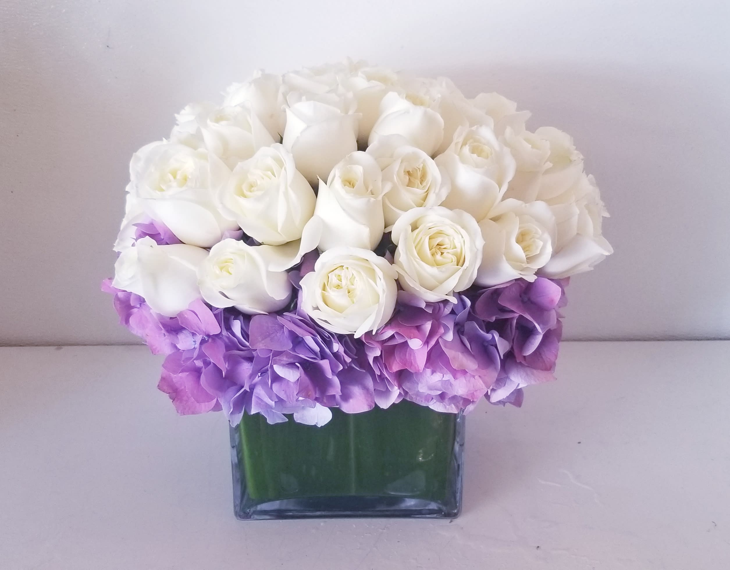 Immaculate - Elegant and clean low and lush arrangement composed of purple hydrangeas surrounded by all white roses.
