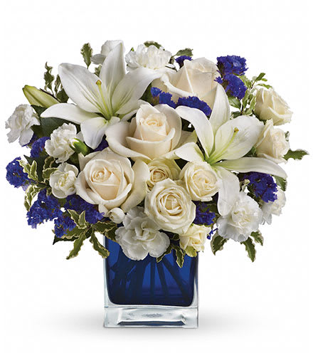 Sapphire Skies Sapphire Skies - Send someone a bit of heaven with this beautiful bouquet. Luxurious crème roses and pure white lilies paint a peaceful picture inside a sapphire blue cube. Crème roses, white asiatic lilies and white miniature carnations are mixed with bursts of purple statice and green pitta negra. Delivered in a glass Cube