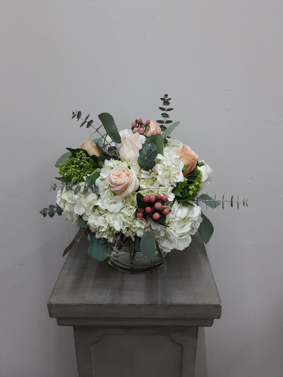 You had me at Hy(drangea) Bouquet - Low arrangement of peach roses, pink hypericum and green hydrangeas on a cloud of white hydrangeas. Eucalyptus adds the perfect touch to this love cylinder arrangement! Such a classy and soft romantic arrangement.  Flowers and colors may vary depending on availability.