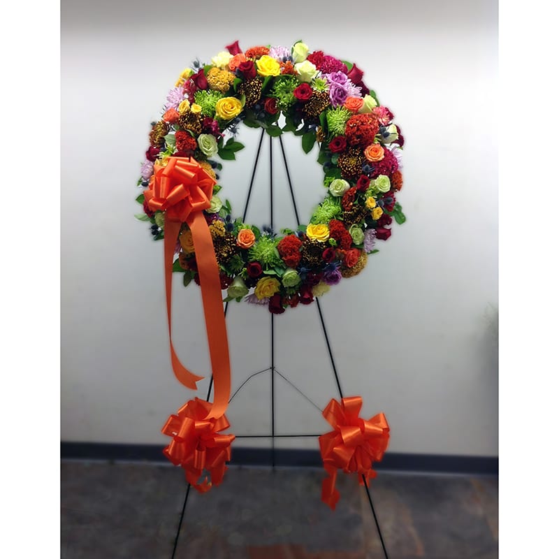 Peaceful Bright - Serene devotion to honor a loved one. This wreath will bright friends and family even in a sorrowful moment. Combination of green spider and brown-golden mums, reds, oranges and yellow roses decorated with orange satin ribbon - accentuated with assorted greenery- in easel.