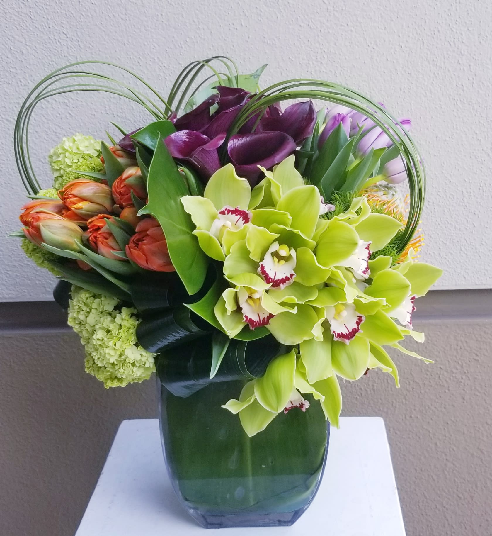 Selena - low colorful exotic arrangement composed of plum mini callas, tulips and cymbidium orchids. It also had folded tropical leaves and grass loops.