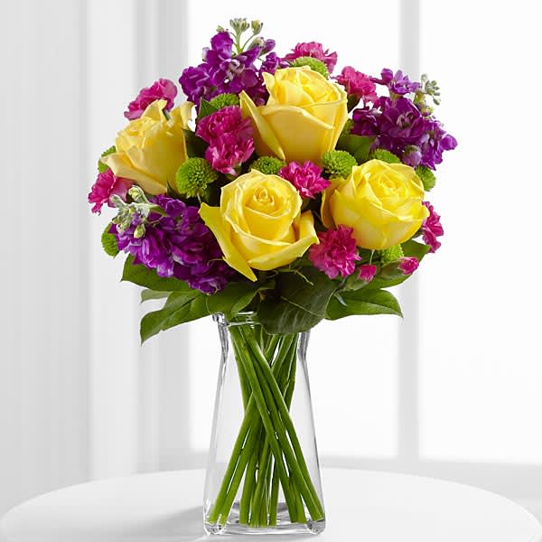 The FTD Happy Times Bouquet - The FTD® Happy Times™ Bouquet employs roses and stock to bring vibrant color and fragrance straight to their door on their special day. Yellow roses purple stock green button poms fuchsia mini carnations and lush greens create a stunning display beautifully arranged in a clear gathered square glass vase to help you convey your happy birthday wishes or send your congratulations.