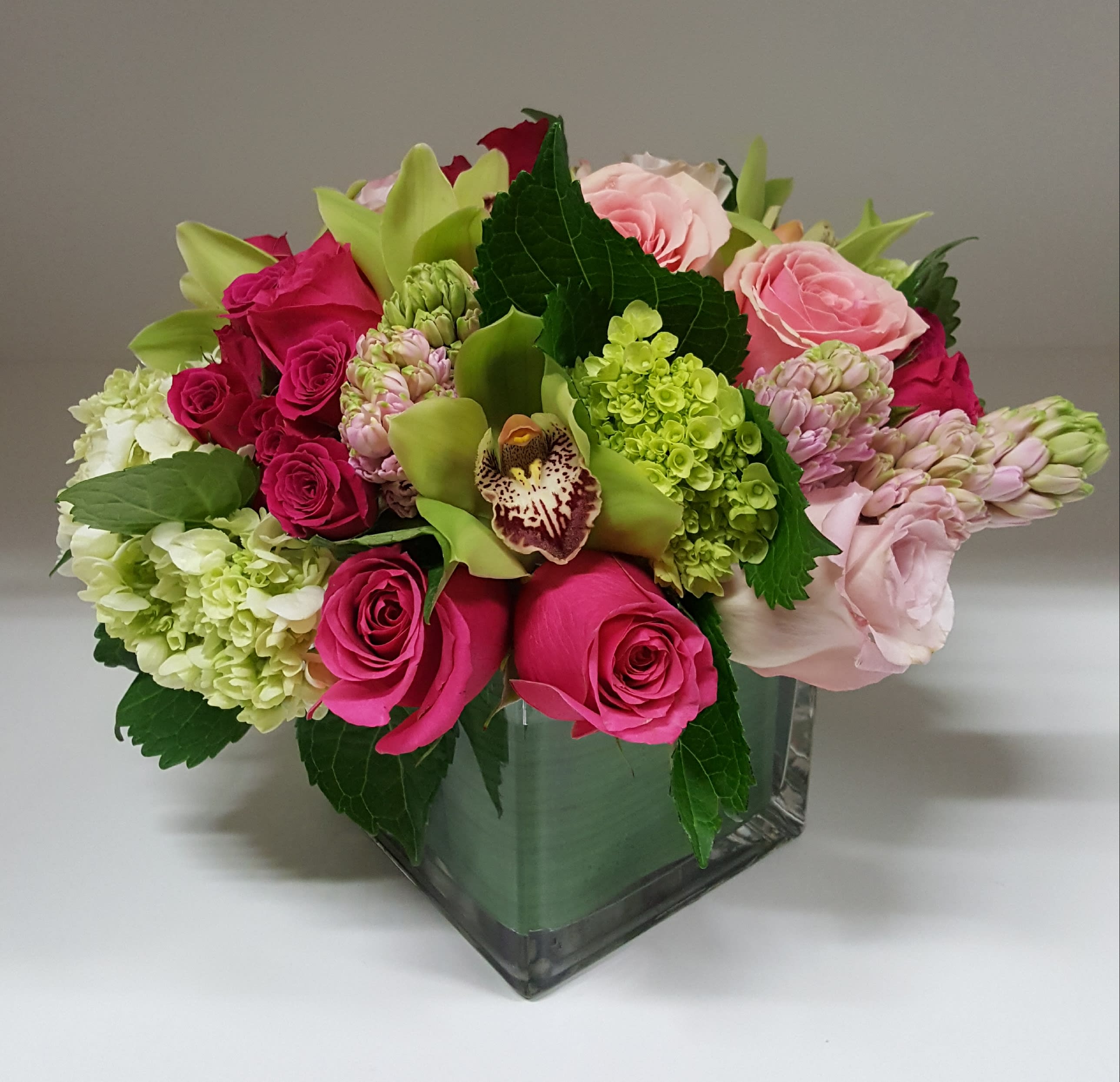 Dragon Fruit - This delightfully vibrant design is created through a contrasting display of fragrant hot pink roses and bright mini green hydrangea alongside with bold green cymbidium orchid blooms set against lighter shades of pink roses and delicate aromatic hyacinth, presented in a 5”x 5” leaf lined clear cube. Approximate dimensions: 11”x 10”