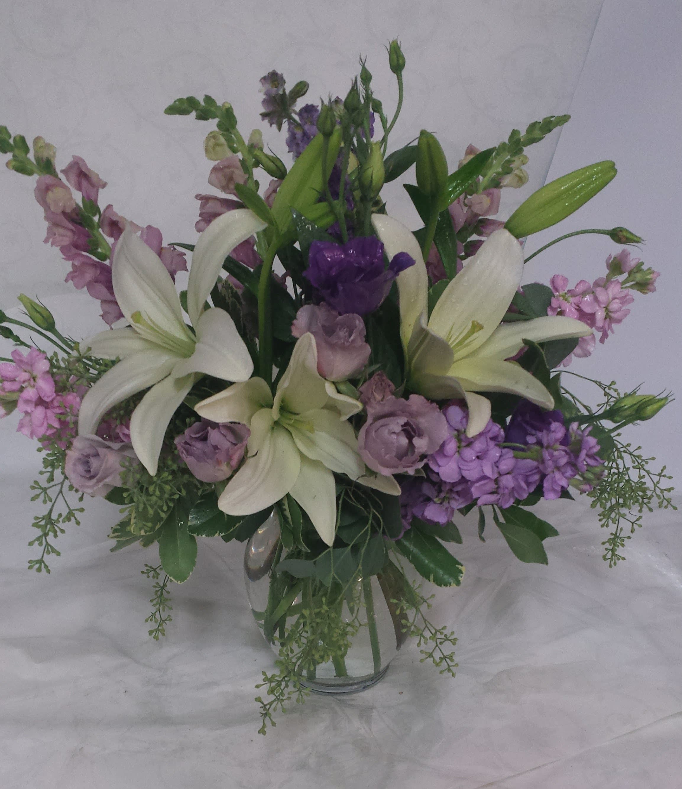 Coolwater - A white and lavender bouquet arranged in a clear glass vase is with  white lilies, lavender stock and coolwater roses. Easy, breezy