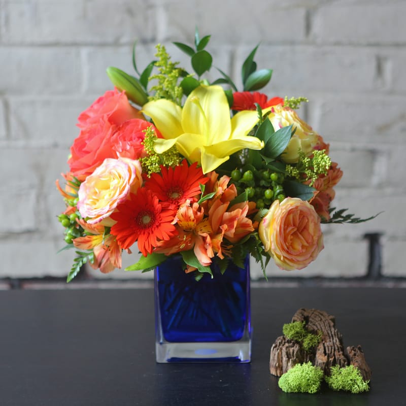 Caribbean Getaway - Stunning shades of orange roses, gerberas, and alstromeria. Complimented by yellow asiatic lilies, solidago, two toned yellow and pink roses and green hypericum berries in a blue glass cube.