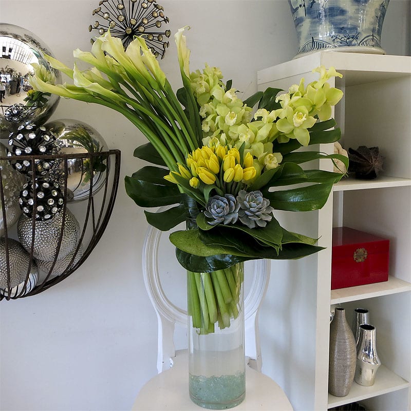 Jaded Yellow Jaded Yellow - Striking arrangement composed of cool, understated colors and clean lines. White calla lilies, pale green cymbidium orchids, and yellow tulips decorated by fanning monstera leaves and pale blue succulent plants.