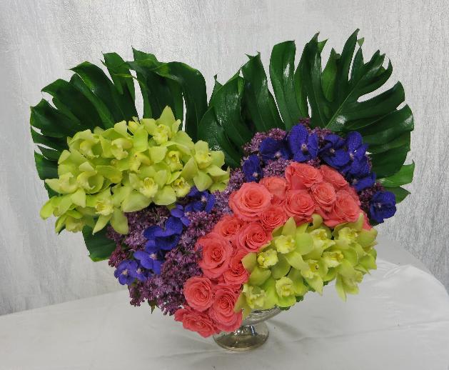 Tropical Heart - An extravagant sweetheart arrangement in a pedestal vase overflowing with two different kinds of Orchids, large Roses, sweet-scented Lilacs, and finished with large Monstera leaves.  *vase subject to availability