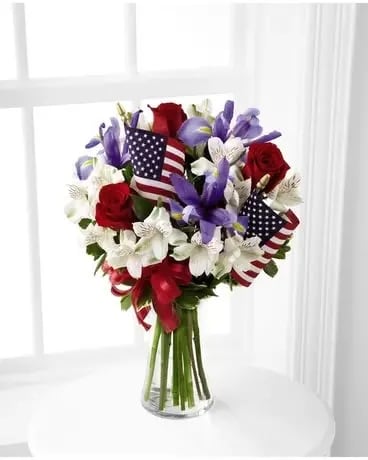 Unity Bouquet - Bright red roses mingle with blue iris, white Peruvian lilies and assorted greens. Accented with two American flags and a red, white and blue ribbon, this stunning bouquet arrives arranged in a clear glass vase for a gorgeous way to celebrate our Independence Day. Standard is approximately 14&quot;W by 18&quot;H Deluxe is approximately 15&quot;W by 19&quot;H Premium is approximately 16&quot;W by 20&quot;H