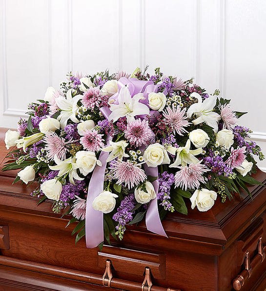 Lavender Half Casket Cover - When we lose someone dear to us, we want to respond with a fitting gesture of respect and admiration. Our half casket cover, crafted with care and artistry by our expert florists with lavender cremones, mums, white roses, and accented with a lavender satin ribbon, is an unforgettable way to commemorate a lifetime of loving devotion.  Half casket cover arrangement of white roses, hybrid lilies and monte casino, lavender cremones, spider mums and stock, purple waxflower and fresh greenery; accented with a lavender satin ribbon Arrangement measures approximately 16&quot;H x 38&quot;L x 26&quot;D Traditionally sent by the immediate family to the funeral home Our florists use only the freshest flowers available, so colors and varieties may vary