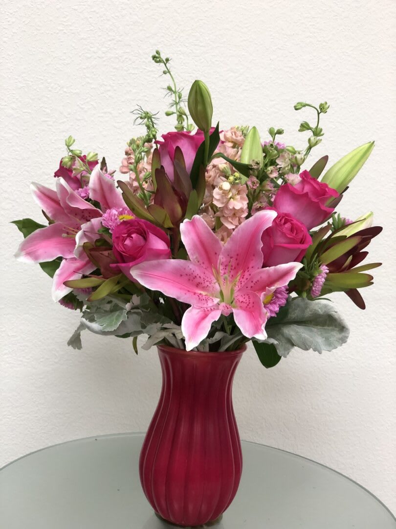 LUSH BRUSH LUSH BRUSH - "A jewel of a gift! Bursting with a lush bouquet of hot pink roses and pink lilies, this breathtaking, hand-blown art pink glass vase has a finish and soothing that’s sure to make her smile! This bouquet features hot pink roses, pink asiatic lilies, purple pink alstroemeria, lavender pink stock, statice, seeded eucalyptus, and leatherleaf fern and dusty miller. Delivered in a hot pink Glass vase. Orientation: All-Around All prices in USD ($)"