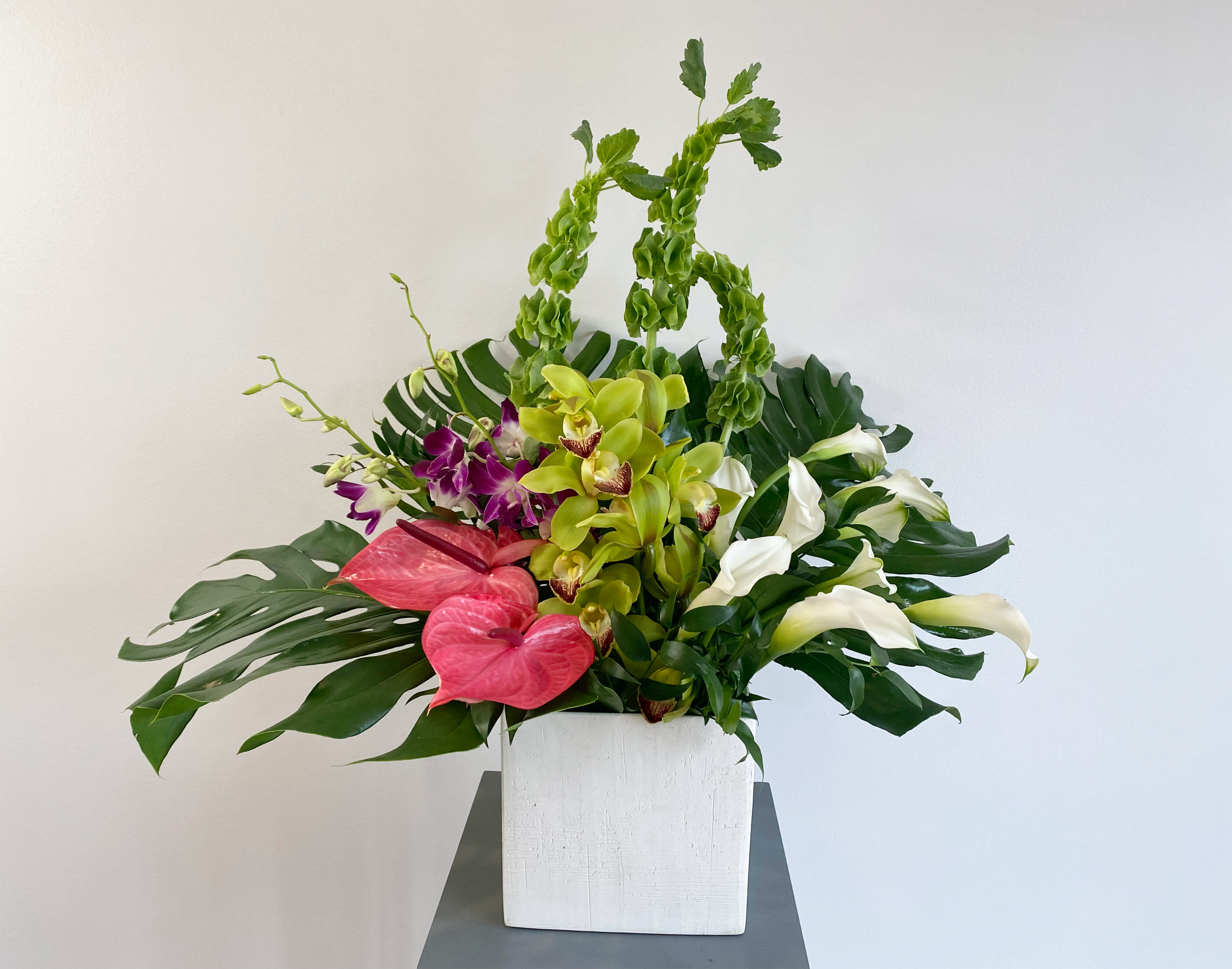 Island Vibez - A tropical knockout. Green Cymbidium, White Cala Lily, and Anthuriums surrounded by huge Monstero Leaves 